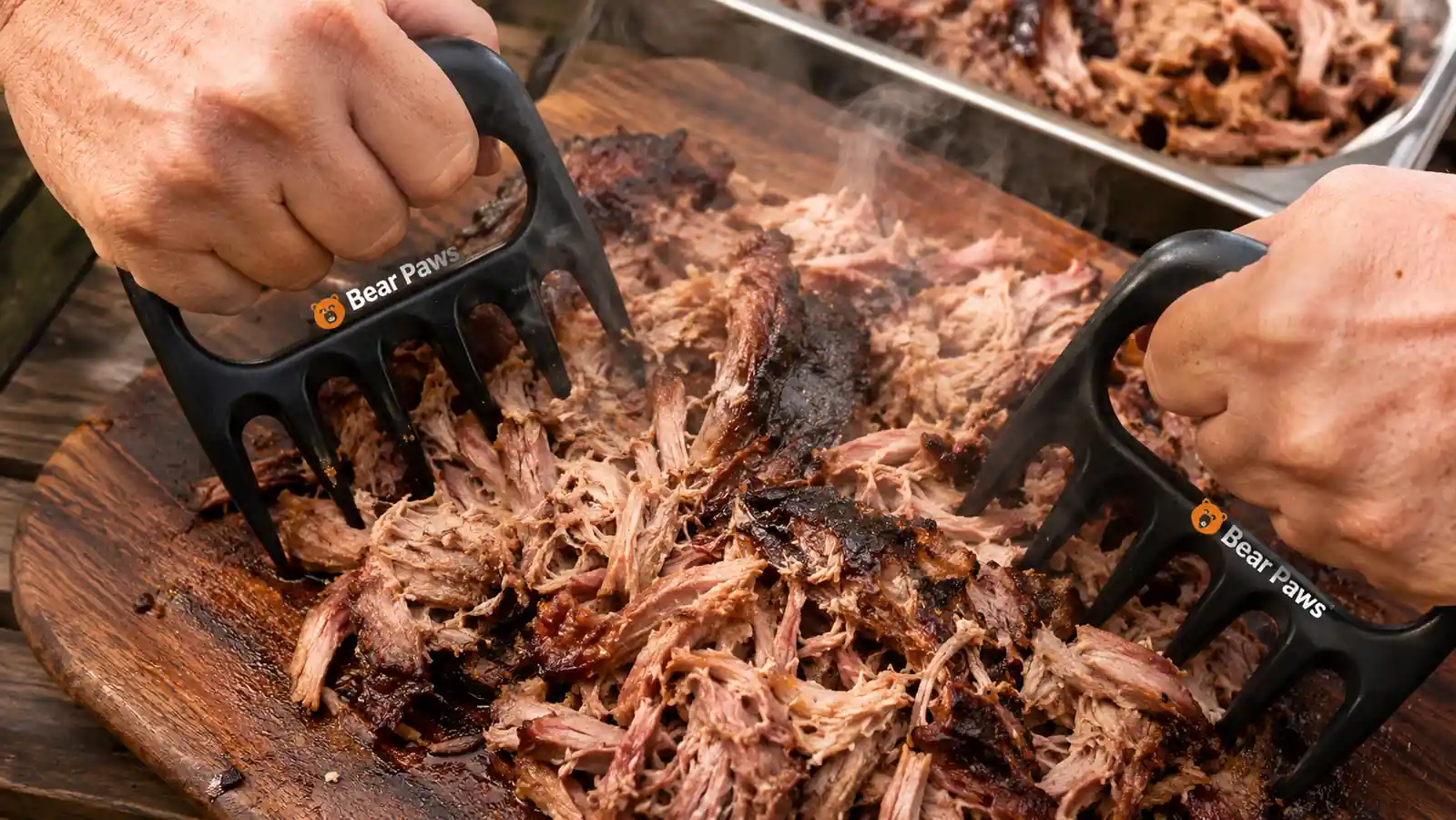Hands using Bear Paw meat claws to shred a pork butt into strands on a cutting board