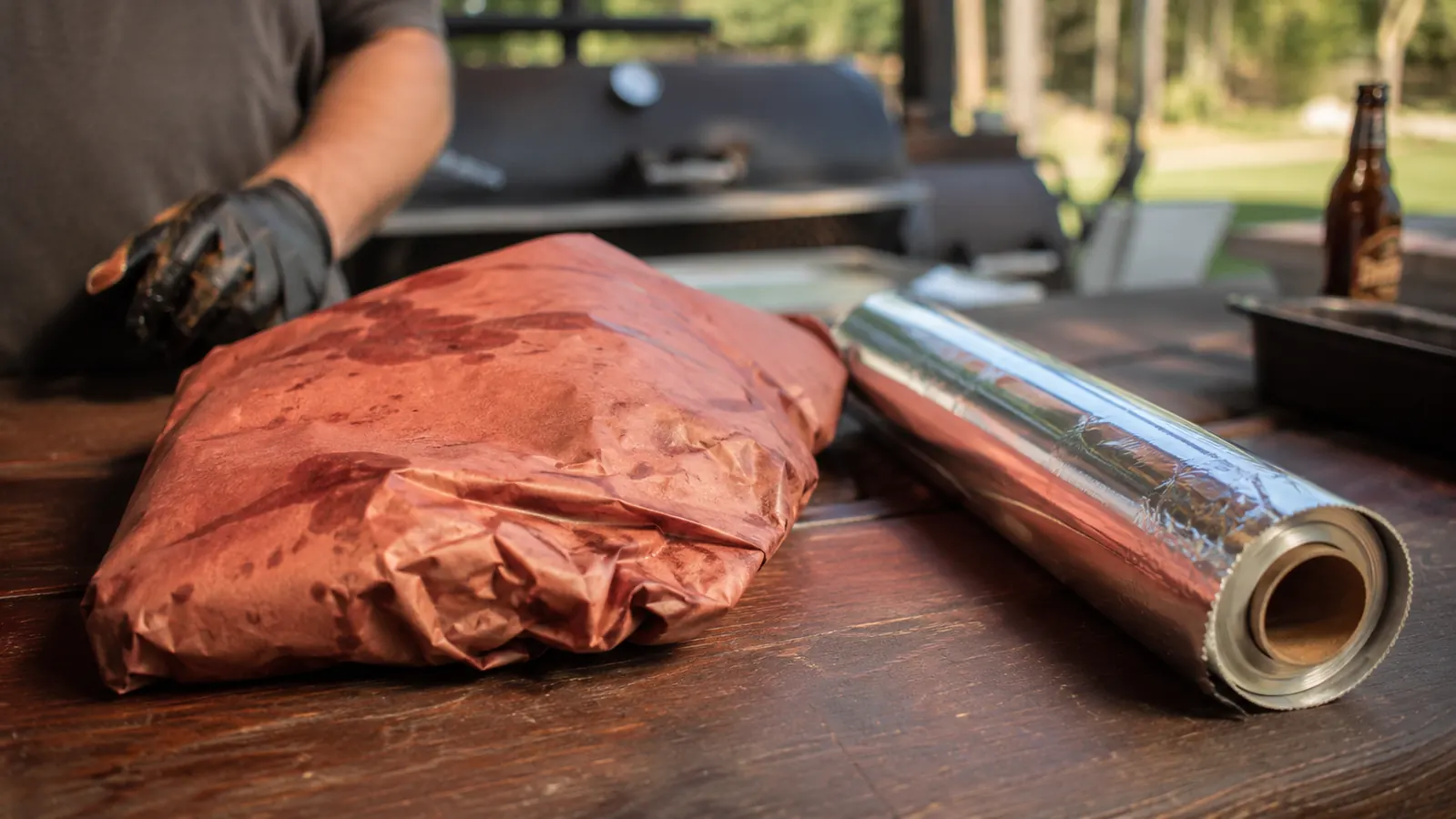 A wrapped brisket resting on a sheet of pink butcher paper next to a roll of aluminum foil on a wooden picnic table