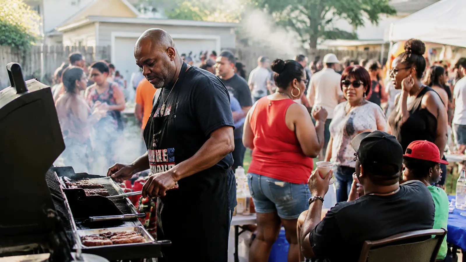 Wide shot of a backyard Fourth of July cookout with a smoking grill and family gathered around picnic tables