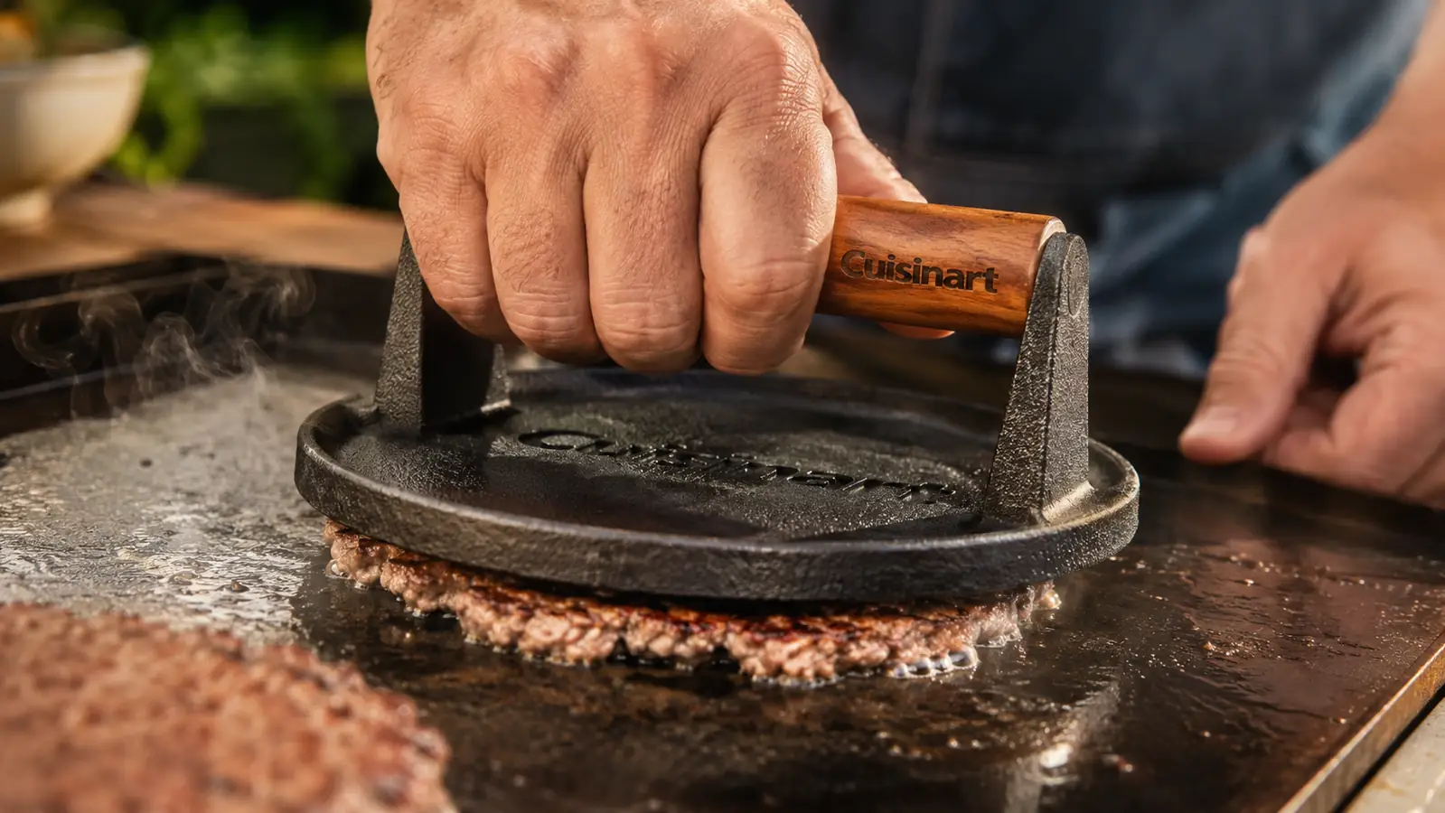 Hand pressing down on a cast iron grill press over a smash burger patty on a griddle