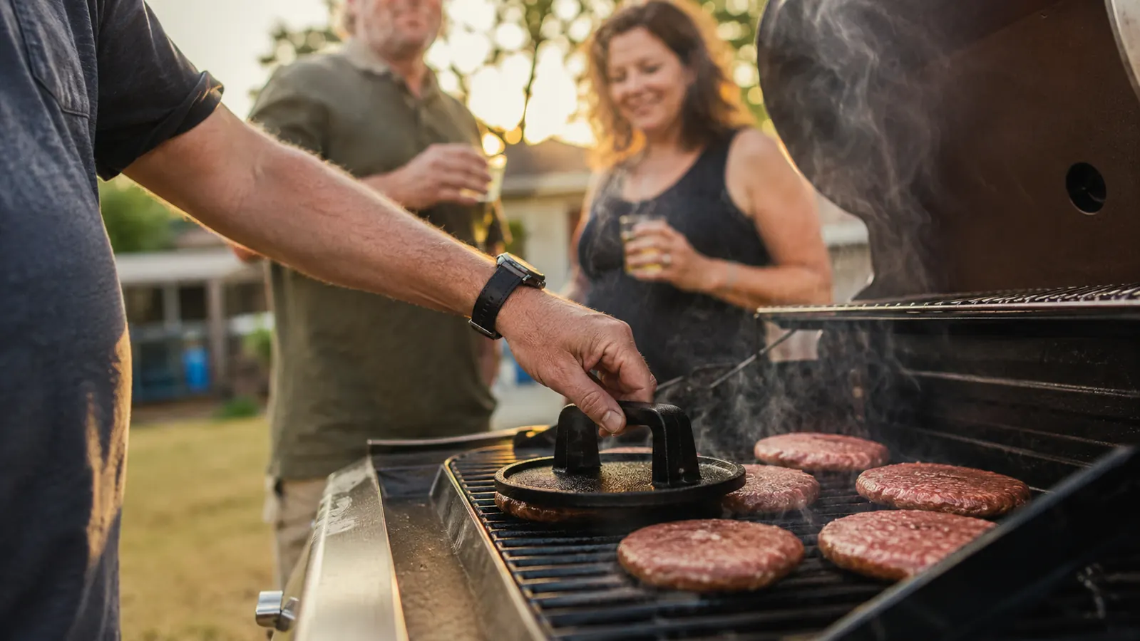 Cast iron grill press sitting on a hot grill grate next to a stack of smash burger patties