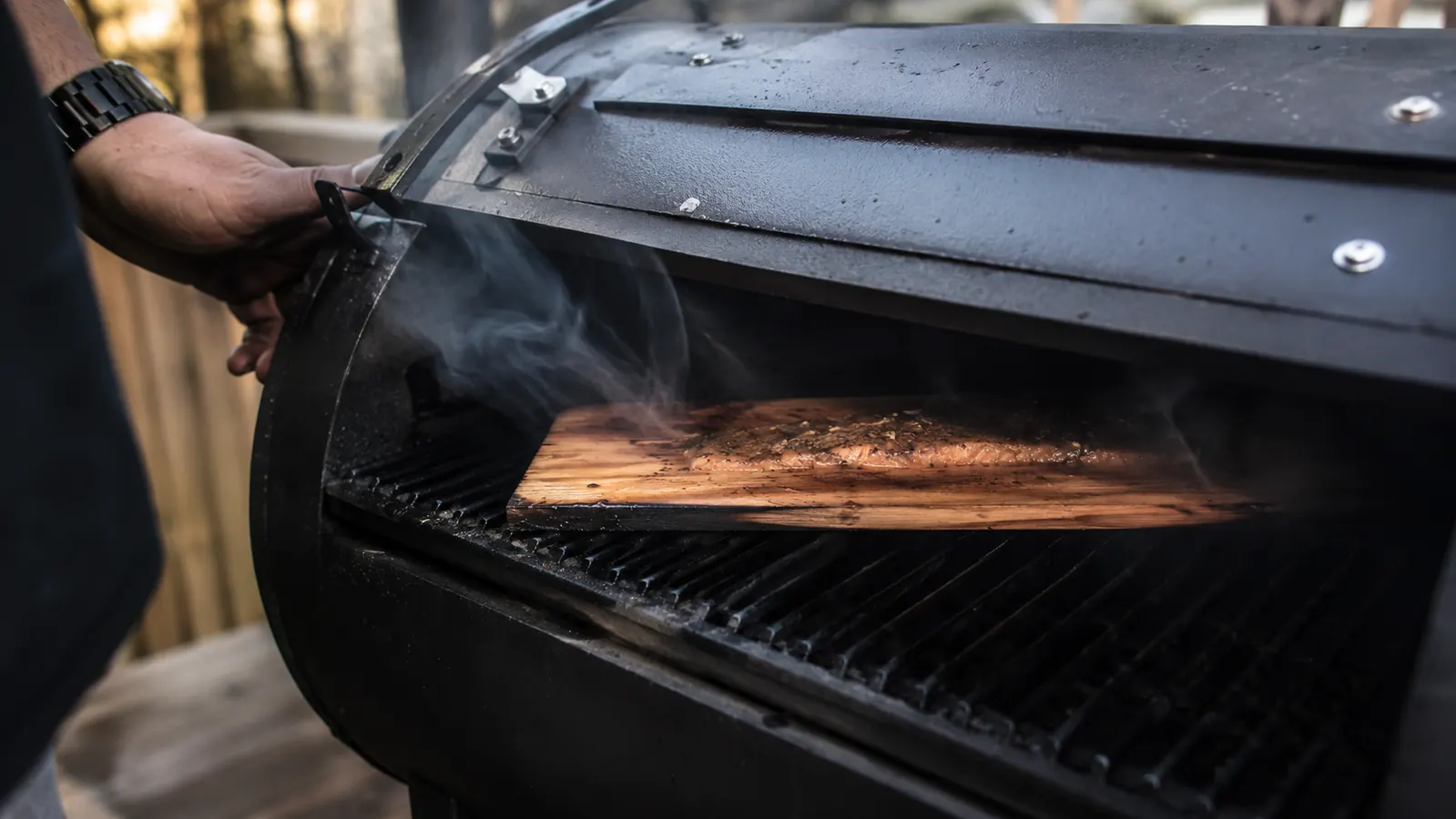 A slightly charred cedar plank smoking on a closed grill grate, smoke curling out the vents