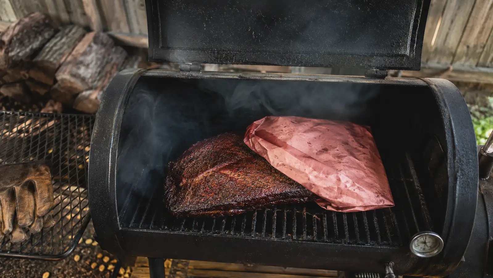 A partially wrapped brisket in pink butcher paper sitting on an offset smoker grate with smoke curling around it