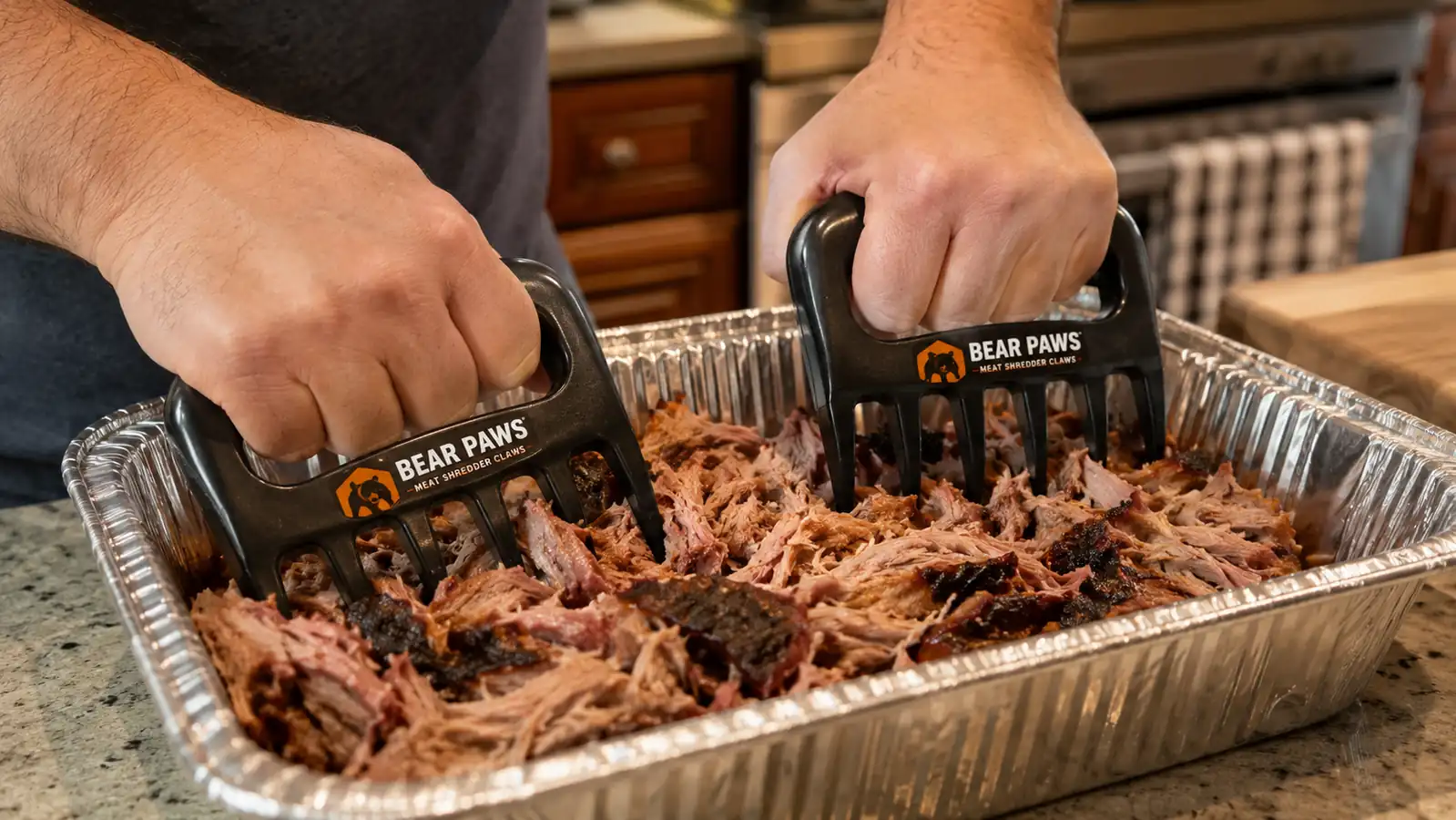 Hands using Bear Paw claws to pull apart a smoked pork shoulder in a foil pan