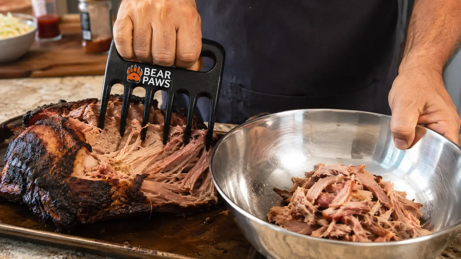 Hand using a Bear Paw meat claw to pull shredded pork into a stainless steel mixing bowl