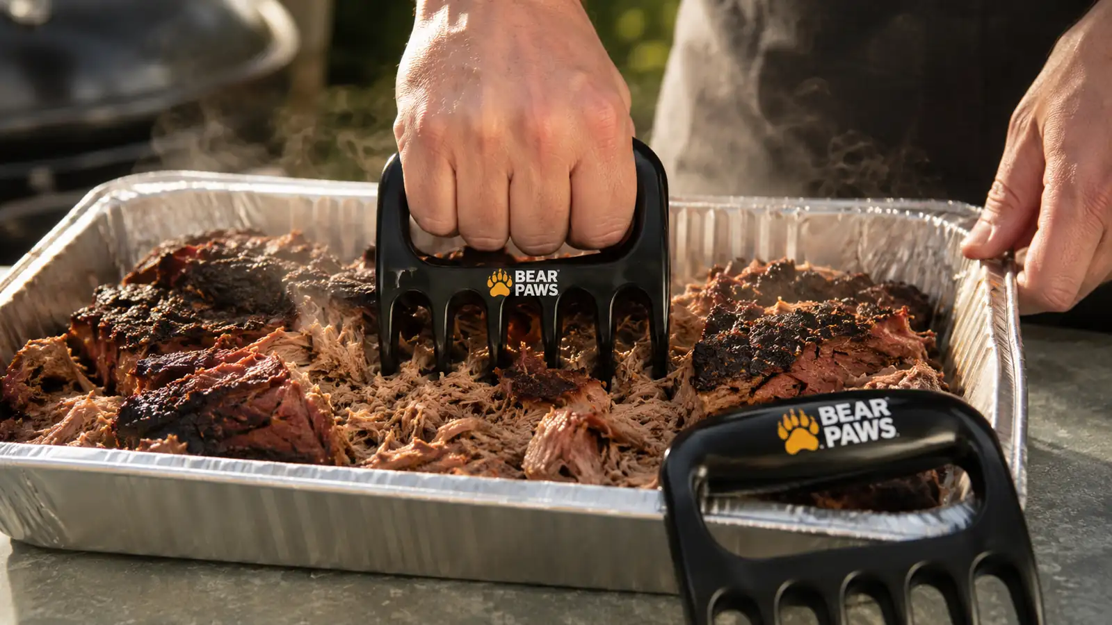 A hand using a Bear Paws meat claw to shred a pork butt in a stainless steel pan